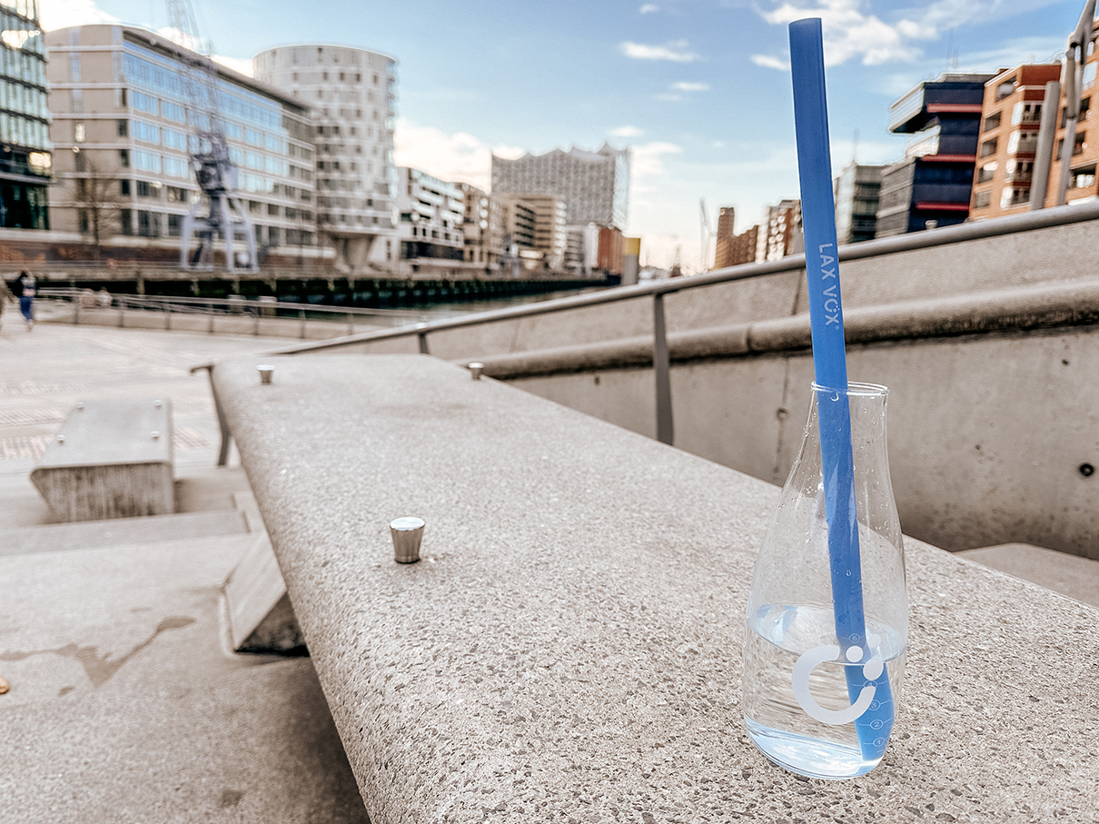 Glasflasche mit einem blauen Trinkhalm auf einer Betonbank in einem modernen Stadtviertel, umgeben von Bürogebäuden und sonnigem Wetter.