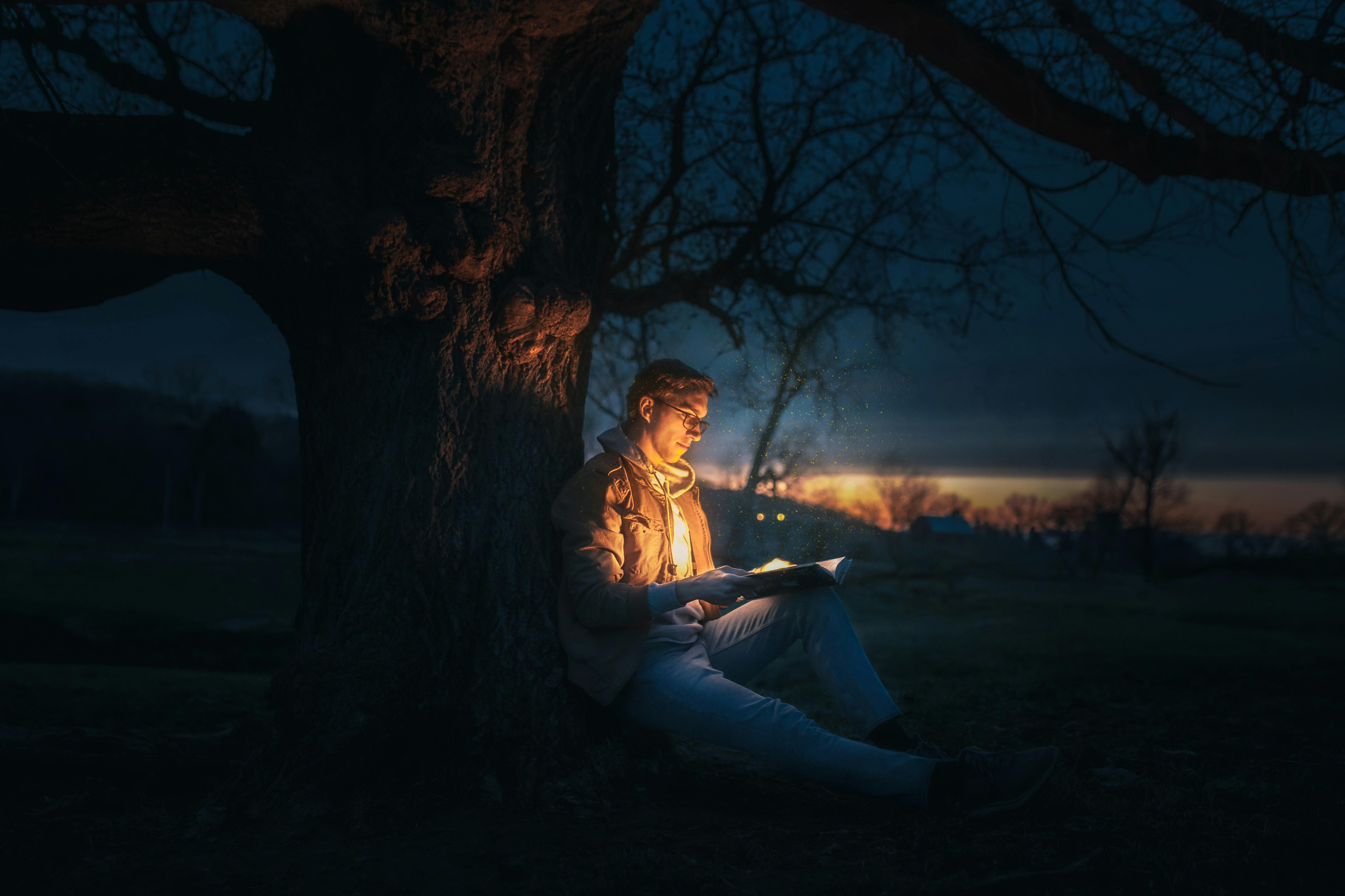 Junger Mann liest ein leuchtendes Buch unter einem Baum bei Nacht, umgeben von Magie.