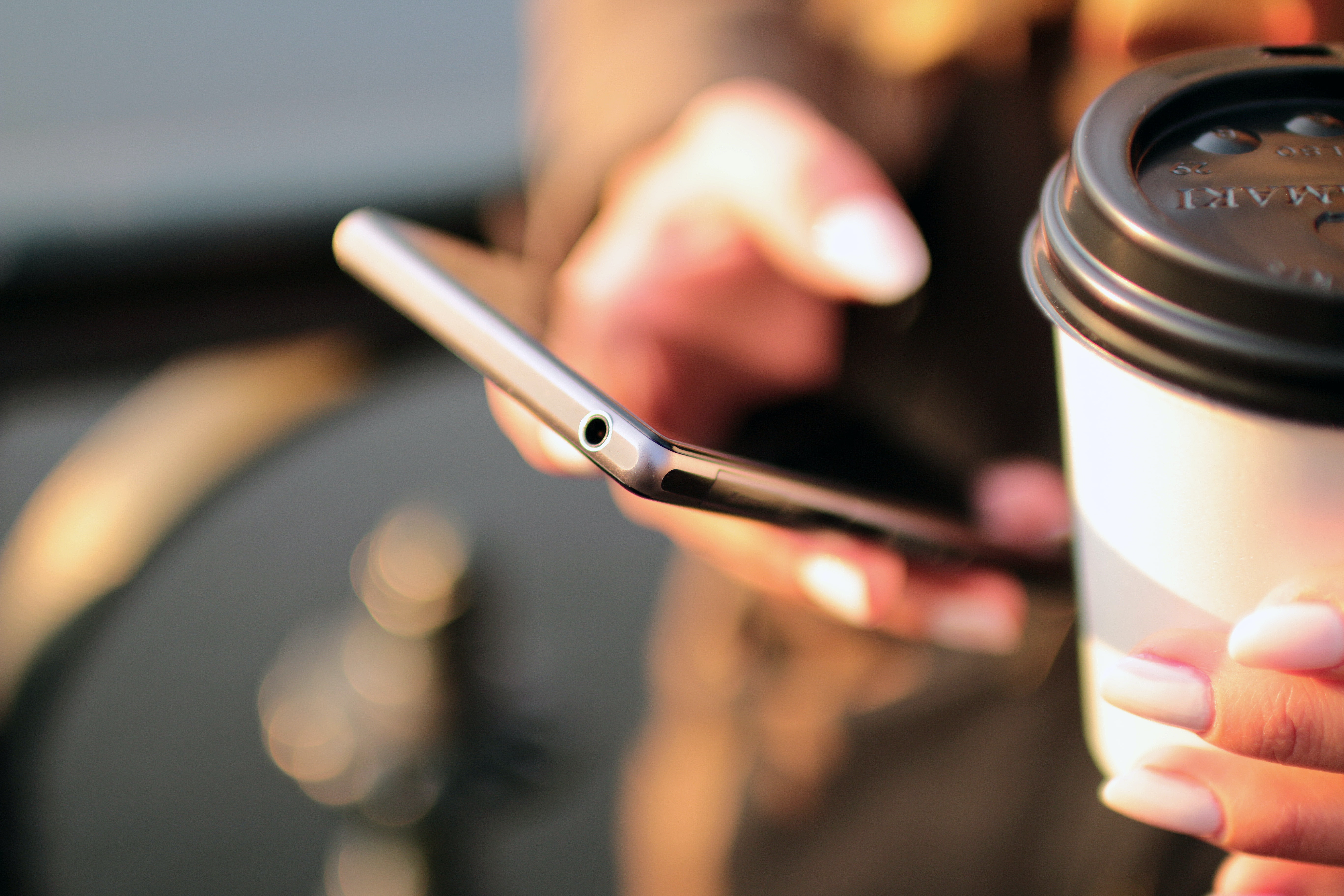 Person hält ein Smartphone und einen Kaffeebecher, beide Hände sind scharf im Fokus.